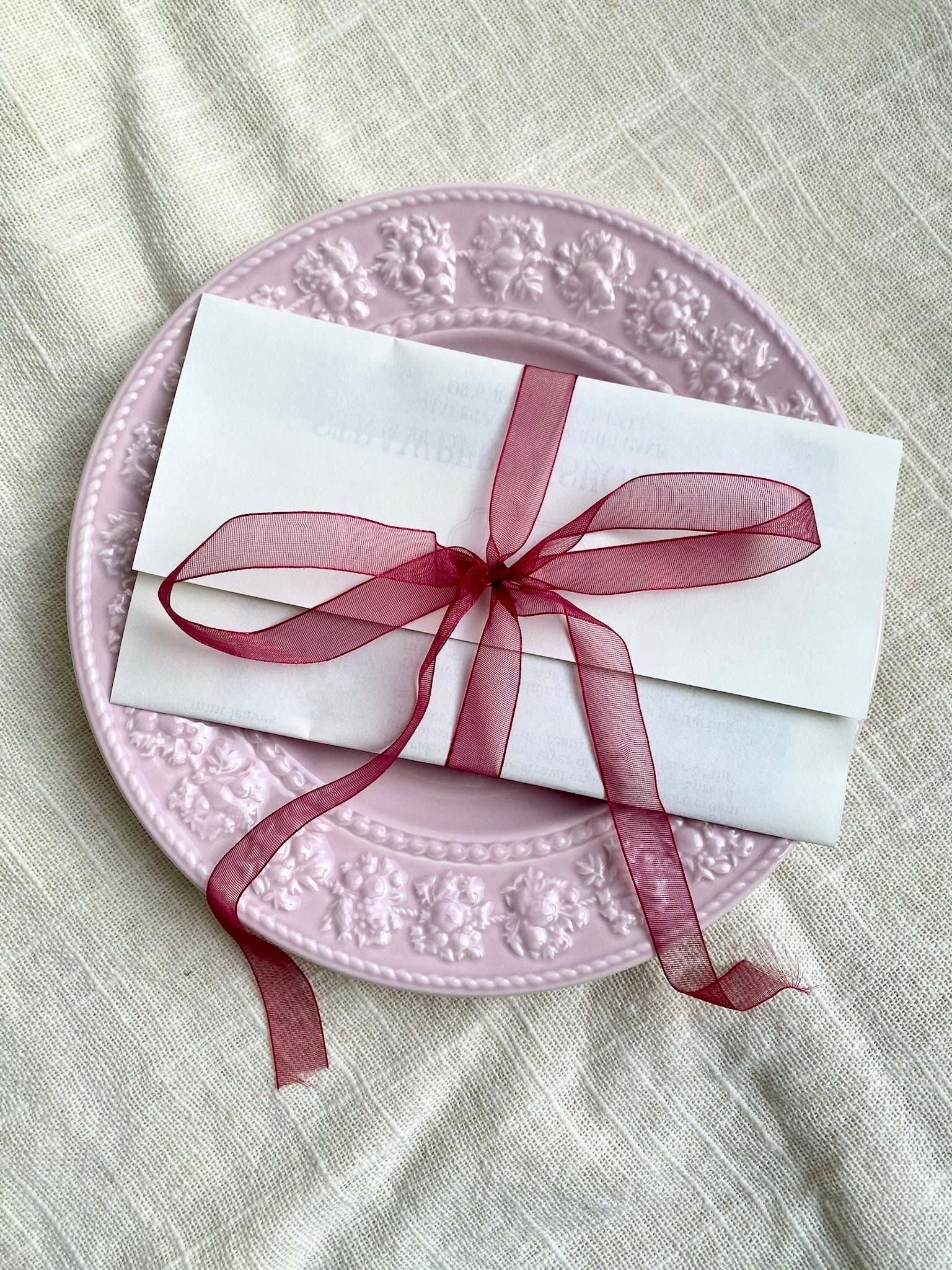 White card with a red ribbon on a pink decorative plate over a textured beige background